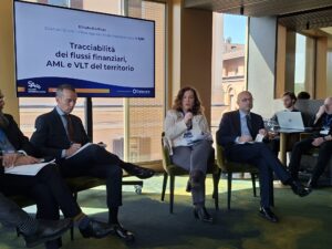 Antiriciclaggio Elisabetta Poso ADM | AGIMEG Panel of professionals in a conference room; a woman speaks into a microphone while others listen, with a large screen behind showing the session title in Italian: 'Tracciabilità dei flussi finanziari, AML e VLT del territorio'