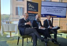 Salone Antiriciclaggio, Pimpinella (APSP): “Lavorare su nuovi modelli di identificazione del giocatore” Three men in suits on a conference panel; the leftmost speaks into a microphone while the others read papers, with a large Italian presentation on screen behind them.