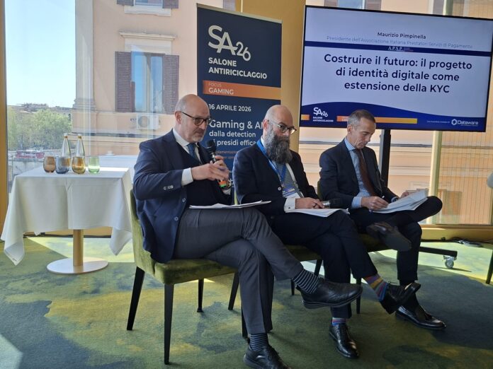 Antiriciclaggio Pimpinella Three men in suits on a conference panel; the leftmost speaks into a microphone while the others read papers, with a large Italian presentation on screen behind them.
