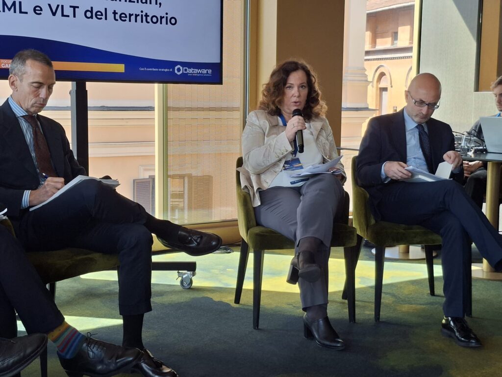 Woman speaks into a microphone during a panel discussion, flanked by two men reviewing papers.