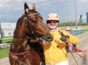 Harness racing driver in an orange suit and helmet giving a thumbs up beside a brown horse at a track.