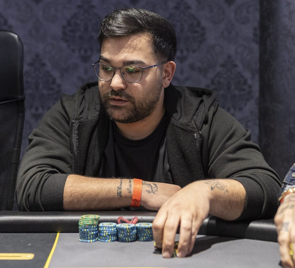Man wearing glasses and a dark hoodie sits at a poker table, stacks of blue chips in front.