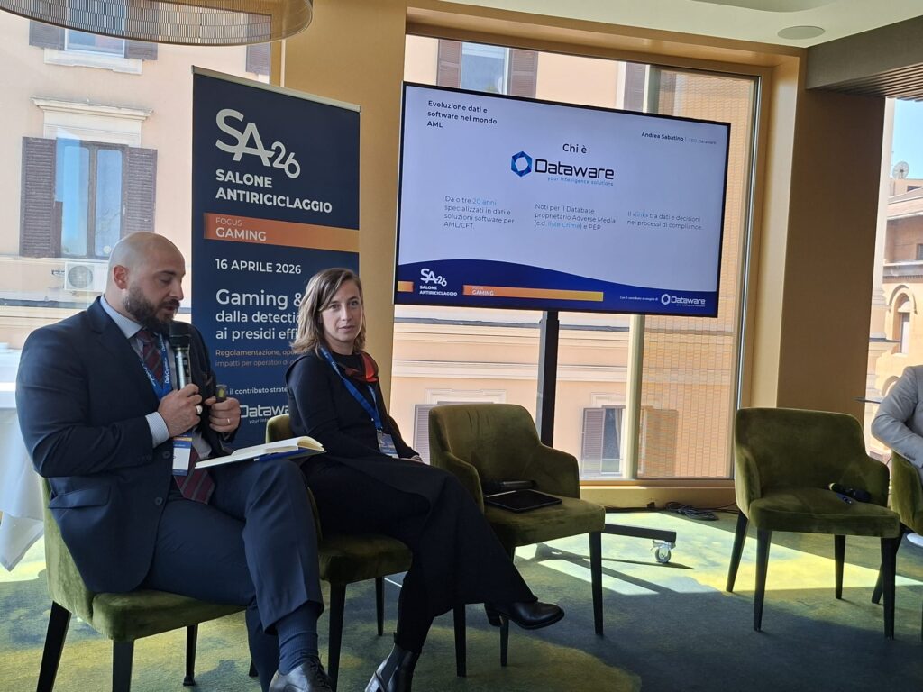 Two presenters sit on green chairs during a conference; a tall banner reads 'SALONE ANTIRICICLAGGIO' beside a large screen displaying a Dataware slide.