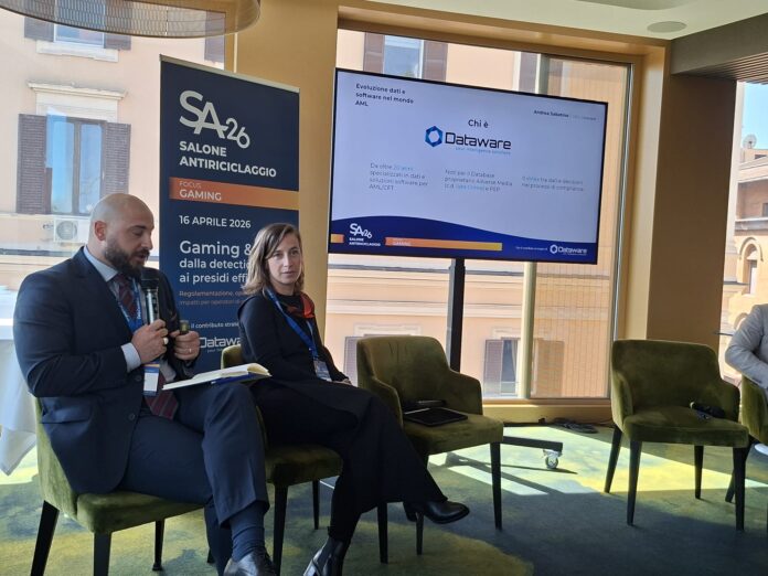 Salone Antiriciclaggio Dataware Two presenters sit on green chairs during a conference; a tall banner reads 'SALONE ANTIRICICLAGGIO' beside a large screen displaying a Dataware slide.