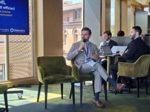 Man in a light gray suit speaks into a microphone during a panel discussion, seated on a green chair with two colleagues at a laptop-filled table nearby.