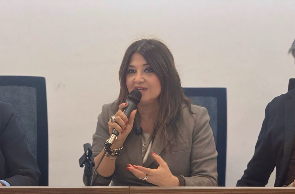Woman in a grey blazer speaking into a handheld microphone at a panel discussion, with hands gesturing.