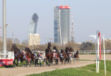 Ippica, Ippodromo Snai San Siro: prima di trotto a maggio dedicata all’allevamento Bigi. Sette prove dalle 14:50 Horses in harness racing teams race on a dirt track with a skyline of modern high-rise buildings in the background.