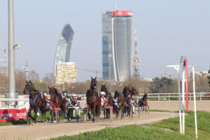 Horses in harness racing teams race on a dirt track with a skyline of modern high-rise buildings in the background.