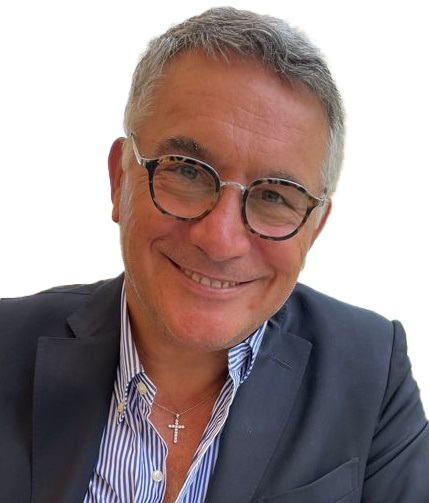 Smiling middle-aged man with gray hair and round glasses, wearing a navy blazer, striped shirt, and a cross necklace.