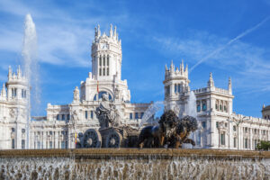 Madrid's Palacio de Cibeles behind the Cibeles Fountain statue of Cybele in a chariot drawn by lions, with water jets and a clear blue sky.