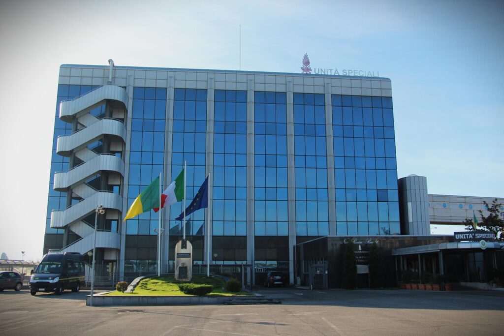 Front view of a modern blue-glass building labeled Unità Speciali with a curved exterior staircase on the left and flagpoles in front.