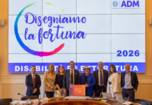 Lotteria Italia 2026, sesta edizione di “Disegniamo la Fortuna con ADM”. Selezionate le opere che saranno riprodotte sui biglietti Group of eight professionals posing and smiling in a conference room in front of a large screen with Italian text and the year 2026.