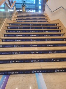Battle of Malta StarYes | AGIMEG Indoor staircase with blue banners reading STARYES and BONUS POKER on each step, leading up to a glass-walled area with plants in gold vases behind.