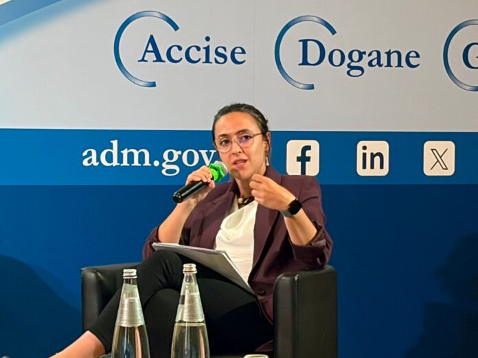 Donata Taddia CIO Woman in a blazer speaks into a handheld microphone on a blue stage backdrop with government logos and social icons.