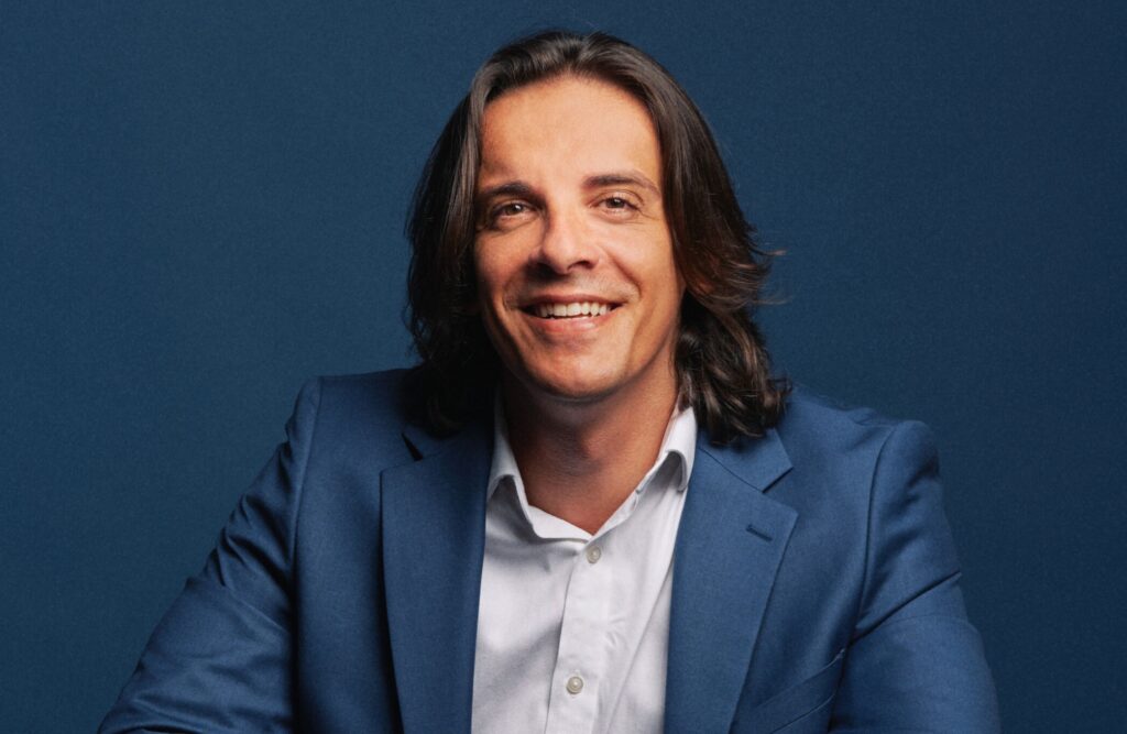 Portrait of a man in a blue suit smiling against a dark blue backdrop, studio portrait.