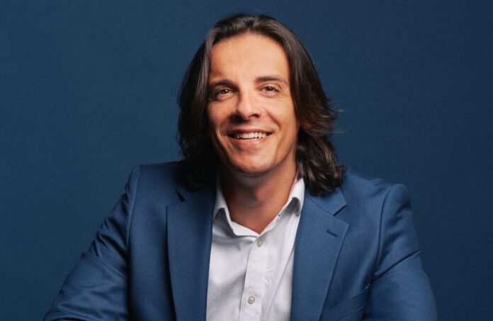 Eman Pulis Portrait of a man in a blue suit smiling against a dark blue backdrop, studio portrait.