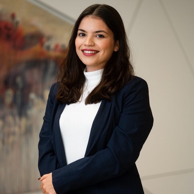 Erika Spiteri Bailey MGA Smiling woman in a navy blazer and white top posing in a bright office hallway.