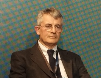 Gallino Agcom | AGIMEG Portrait of an older man in a dark suit and tie, wearing glasses, seated against a teal patterned backdrop.