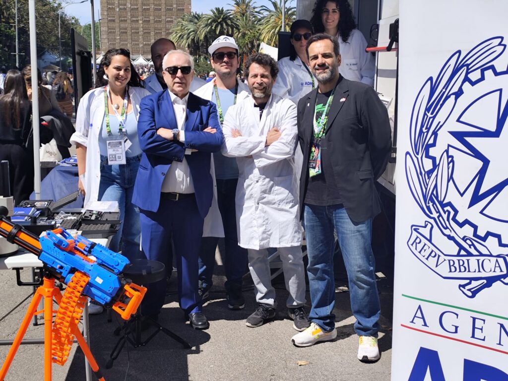 Group of people at an outdoor science event booth, some in lab coats, with orange and blue toy blasters on display in the foreground.