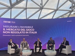 Gioco illegale in Italia convegno | AGIMEG Panel discussion with five men in business attire seated on white chairs on a stage with a large purple banner about measuring the invisible unregulated gambling market in Italy.