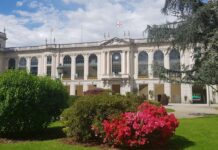 Ippica, è l’ora del “Derby Day”: martedì 2 giugno all’Ippodromo Snai San Siro giornata di grande galoppo, musica e intrattenimento Historic white neoclassical building with large arched windows, surrounded by green lawns and pink flowering bushes under a partly cloudy sky