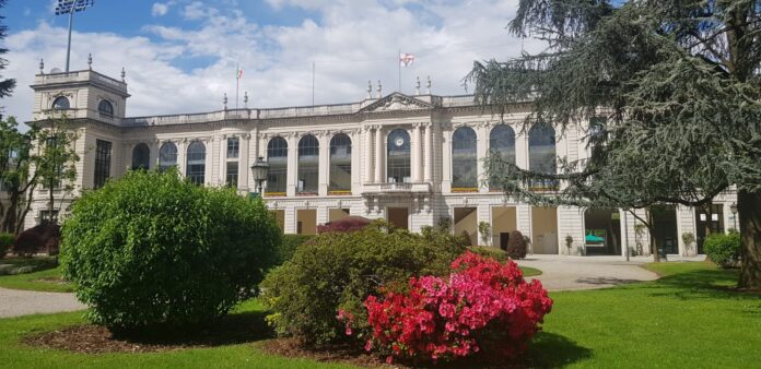 IPPODROMO SNAI SAN SIRO Historic white neoclassical building with large arched windows, surrounded by green lawns and pink flowering bushes under a partly cloudy sky