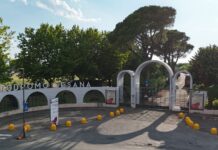 Ippica, Ippodromo Snai Sesana: giovedì riunione di trotto, sette corse a partire dalle 15:40 Entrance gate and curved arches at a walled property with large white letters, facing a road lined with yellow bollards and trees behind.
