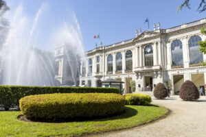 Ippodromo Snai San Siro_Area Fontana | AGIMEG Elegant white neoclassical building with large arched windows and a central fountain spraying water, flagpoles above, manicured shrubs in foreground