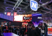 Novomatic si rafforza sul mercato balcanico con nuovi prodotti al Belgrade Future Gaming 2026 Large 'fazi' signage hangs above a crowded exhibition booth with purple neon hex lights and big screens overhead.