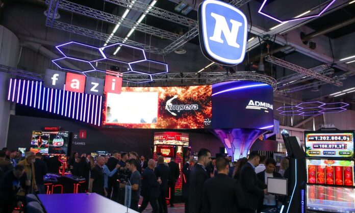 Novomatic Balcani Large 'fazi' signage hangs above a crowded exhibition booth with purple neon hex lights and big screens overhead.