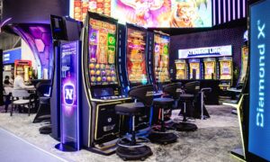 Novomatic mercato Balcani | AGIMEG Row of glowing slot machines with colorful reels and stools at a casino exhibition booth in an expo hall with neon accents and branding.