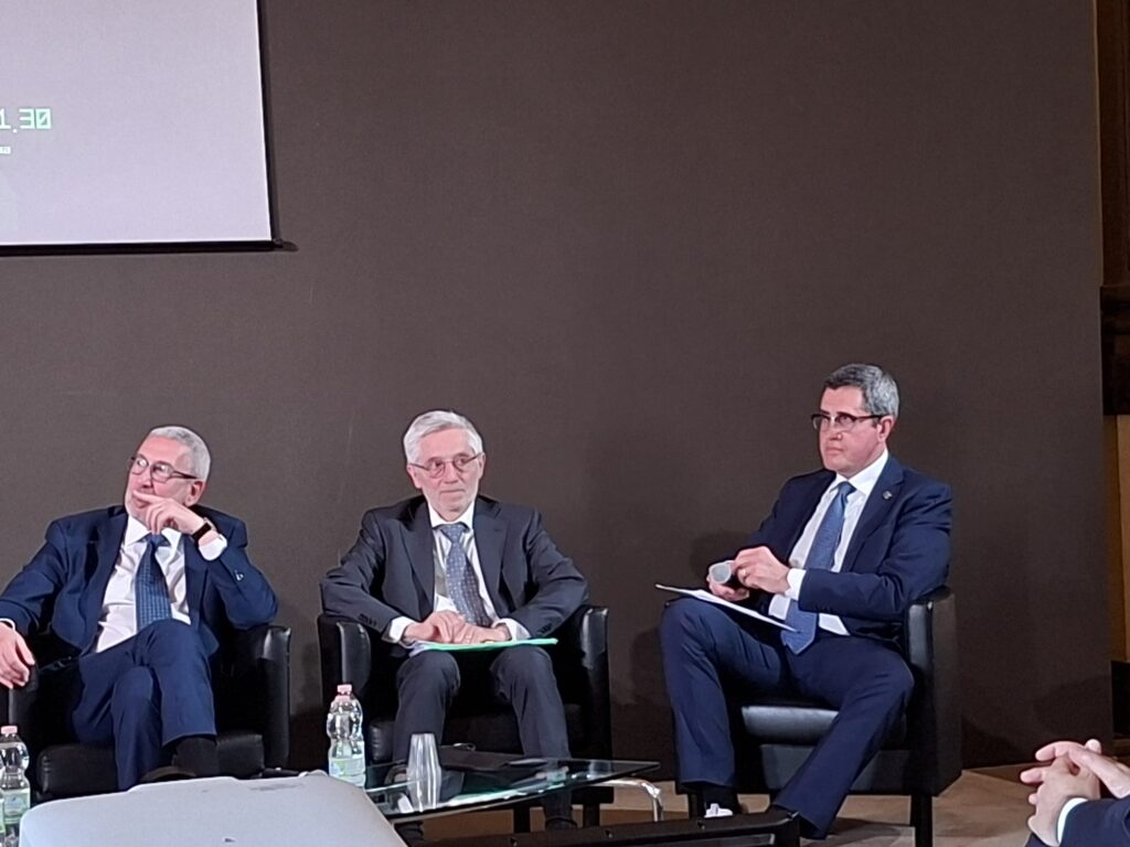 Three men in dark suits seated on a panel stage, engaged in a discussion at a conference.
