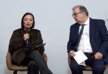 Webinar Gioco responsabile, Siani (Serviceplan Italia): “Serve una nuova comunicazione orientata alla prevenzione” Woman speaks into a handheld microphone while seated, holding a tablet; man in a suit looks on, holding papers in a discussion setting.