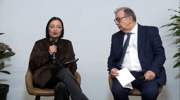 Stefania Siani CEO e CCO Serviceplan Italia presidente ADCI Woman speaks into a handheld microphone while seated, holding a tablet; man in a suit looks on, holding papers in a discussion setting.
