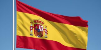 Scommesse illegali e tutela dei minori: la Spagna vara un nuovo piano di azione Spanish flag waving in the wind against a clear blue sky, showing red-yellow-red stripes and the national coat of arms in the center of the yellow band.