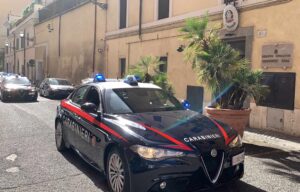 Carabinieri Roma spaccio bisca | AGIMEG Police cars with blue lights parked on a sunlit narrow street outside a beige building in Rome.