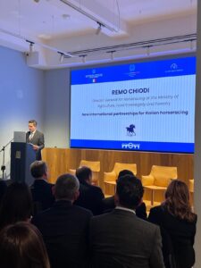 Chiodi Masaf | AGIMEG Speaker at a podium presenting to an audience; large screen shows a slide reading 'Remo Chiodi' and 'New international partnerships for Italian horseracing'.