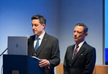 Ippica italiana protagonista a Londra, Chiodi (MASAF): “Rilancio del settore è una sfida globale” Two men in dark suits at a lectern during a formal presentation; one speaks into a microphone beside an HP laptop, the other stands nearby on stage in a conference setting.