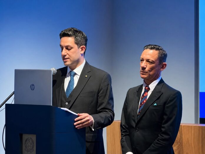 Chiodi Masaf ippica Two men in dark suits at a lectern during a formal presentation; one speaks into a microphone beside an HP laptop, the other stands nearby on stage in a conference setting.