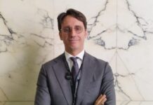 Scommesse, Fiumara (AD GBO – Gruppo Lottomatica): “Finale di stagione calcistica molto positivo grazie ad una offerta innovativa, con la quale puntiamo ad essere protagonisti anche per i Mondiali di Calcio” Man in a gray suit and tie stands against a marble wall, arms crossed, wearing a lanyard with a badge and a clip-on microphone.