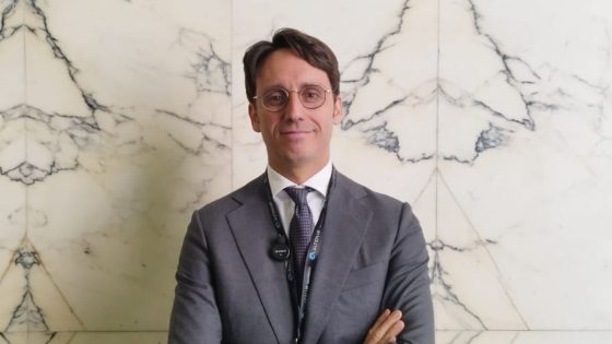 Fiumara Lottomatica Man in a gray suit and tie stands against a marble wall, arms crossed, wearing a lanyard with a badge and a clip-on microphone.