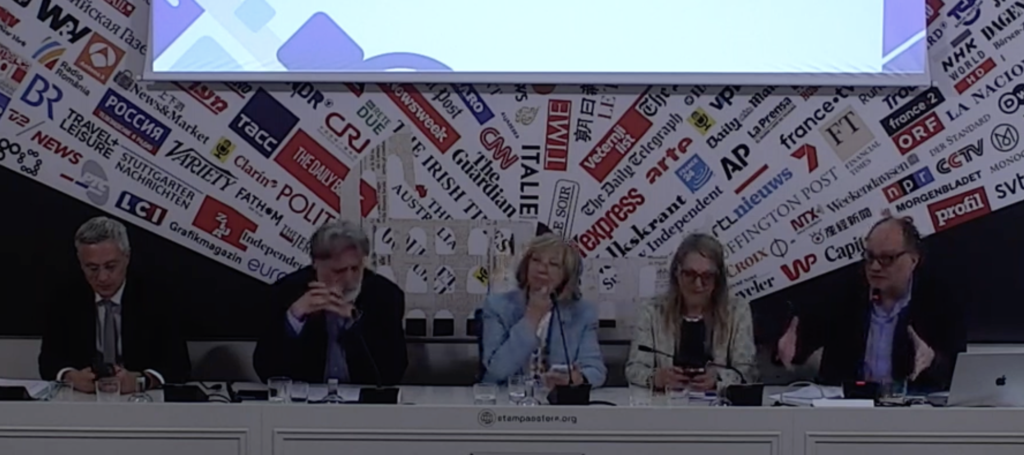 Five panelists seated at a long white desk during a press conference, with a backdrop of various news logos and a projector screen above them.