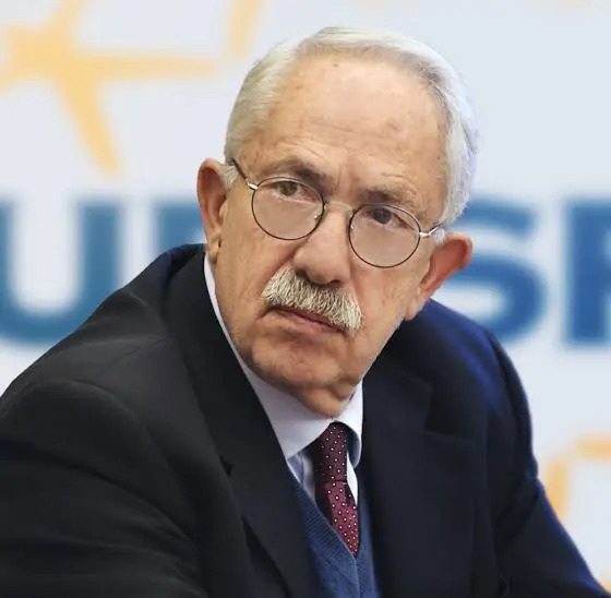 Gianmaria Fara Eurispes | AGIMEG Portrait of an older man with white hair and a mustache, wearing round glasses, a dark suit, light blue shirt, and maroon tie, looking to the left against a blurred backdrop.