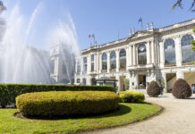 Ippica: l’Ippodromo Snai San Siro si tinge di rosa per le 117^ Oaks d’Italia-Tattersalls White neoclassical building with large arched windows and flagpoles, a dramatic fountain on the left, and well-manicured hedges in the foreground.