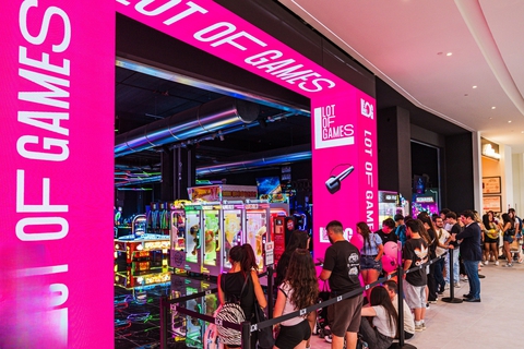 LOG_ENTRANCE | AGIMEG Line of people waiting to play arcade games inside a bright pink gaming booth in a mall.