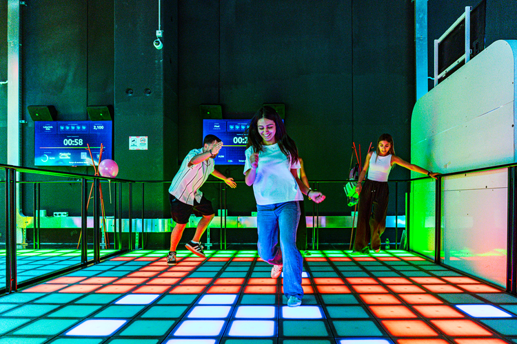 LOG_OPENING2 | AGIMEG Three friends dance on a glowing multicolored LED floor in a dark arcade/club, with screens and equipment visible in the background.