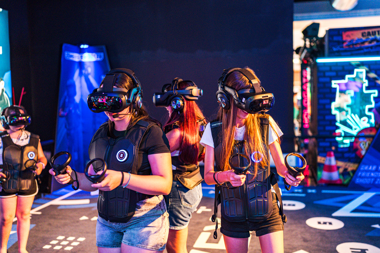 LOG_OPENING3 | AGIMEG Group of people wearing VR headsets and vests in a neon-lit arcade, each holding motion controllers.