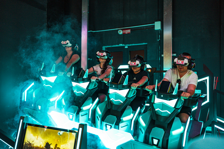 LOG_OPENING4 | AGIMEG Four people wearing VR headsets sit in glowing neon racing simulators at a dimly lit arcade, with cyan lights and smoke.