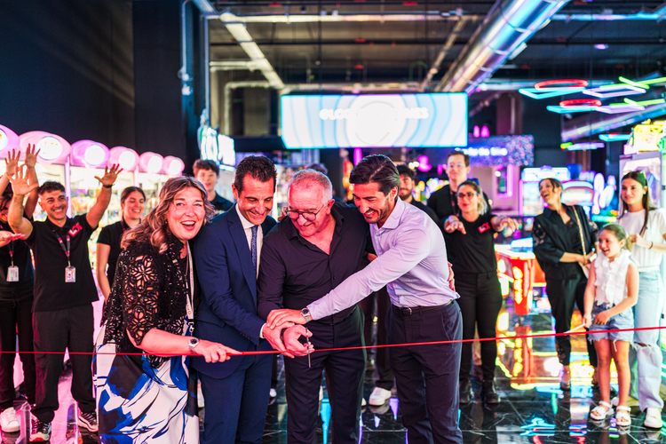 LOG_OPENING5 | AGIMEG Group of people cutting a red ribbon at a ribbon-cutting ceremony in a neon arcade setting with onlookers cheering nearby, bright neon lights in the background.