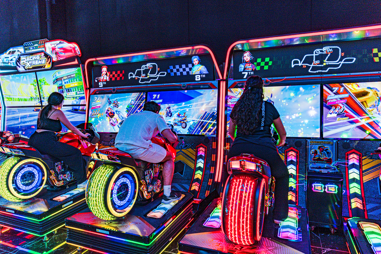 LOG_OPENING6 | AGIMEG Three players on neon-lit motorcycle arcade bikes in a bright arcade, each screen showing racing action.